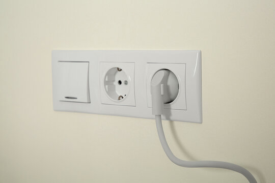 Power sockets with inserted plug and light switch on white wall indoors