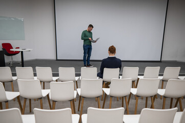 A red-haired Caucasian businesswoman sits in the front row of an empty conference room. Bearded man giving a lecture. 