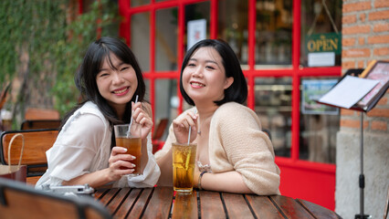 Two happy Asian female friends are enjoying their drinks, sitting at an outdoor table in a cafe.