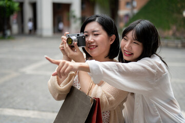 Two happy Asian female friends are strolling around the city, using a film camera to take a picture.