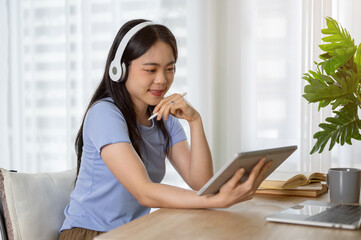 A pretty Asian woman listening to music through her headphones while using her tablet at a table.