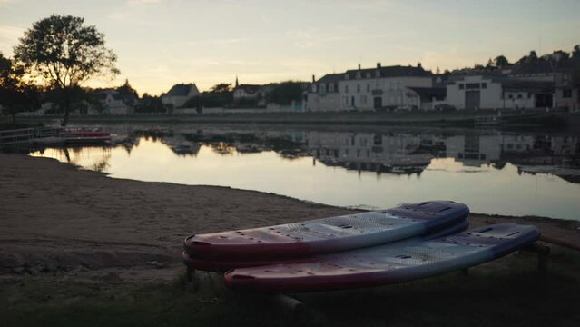 Paddle boards sit idle next to a peaceful river and small village as evening closes in