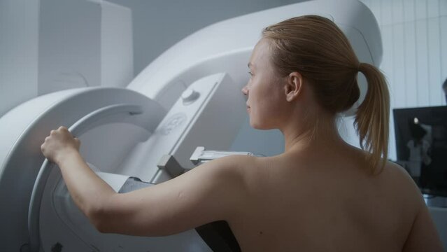 Caucasian adult woman stands topless in clinic radiology room. Female patient undergoing mammography screening diagnostic using digital mammograph. Breast cancer prevention. Modern hospital equipment.