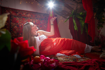 Beautiful European girl looking like Arab woman in red room with rich fabrics and carpets in sultan...