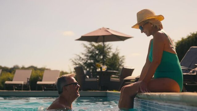 Senior retired couple on holiday with man swimming to woman sitting on edge of hotel pool - shot in slow motion