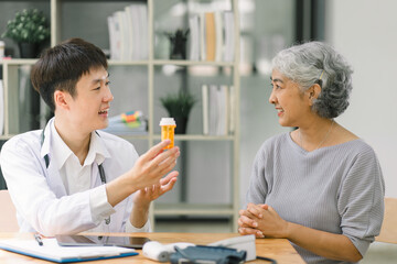 Obraz premium Smiling young asian male doctor explaining the bottle of pills to senior woman patient in modern clinic hospital. Medical healthcare concept.