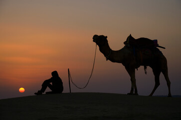A man sitting with camel along with Sunset at Khuri sand dunes, Jaisalmer, Rajasthan, India, Asia.