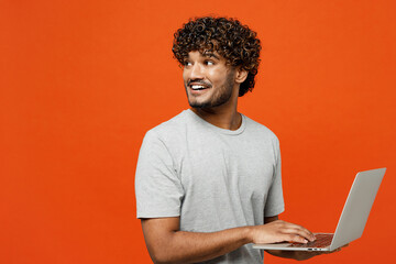Side view young smiling happy Indian man wears t-shirt casual clothes hold use work on laptop pc computer look aside on area isolated on orange red color background studio portrait. Lifestyle concept. - Powered by Adobe