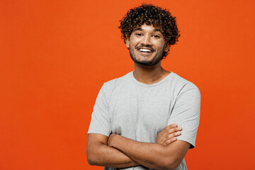 Young smiling happy confident cheerful Indian man he wearing t-shirt casual clothes hold hands crossed folded loking camera isolated on orange red color background studio portrait. Lifestyle concept.