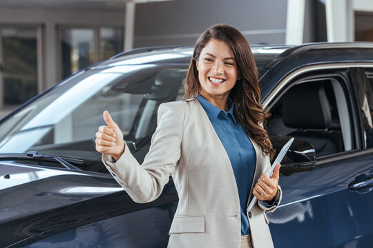 Professional car dealer posing proudly at auto showroom, smiling to the camera, showing thumb up. Happy saleswoman working at automobile dealership, copy space. Professionalism, management concept