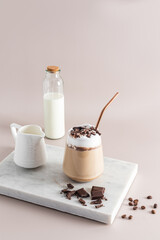 A glass of coffee latte or cappuccino with chocolate wedges stands on a marble white podium with a bottle and a jug of milk. Vertical front view.