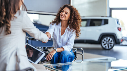 Happy woman buying a car and closing the deal with a handshake with the saleswoman at the...