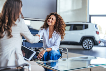 Happy woman buying a car and closing the deal with a handshake with the saleswoman at the dealership. Smiling car saleswoman discussing a contract with a female customer. - obrazy, fototapety, plakaty