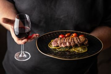 A man in black holds a roasted duck breast fillet on caramelized apples and a glass of red wine on a black background