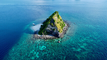 A small rocky uninhabited island in the ocean and a boat on the shore. Aerial view of an uninhabited island in the blue sea, a coral reef around and a small motor boat off the shore.