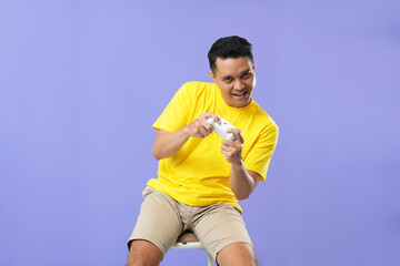 Fototapeta na wymiar Photo of cheerful Asian man playing video game with joystick wearing yellow t-shirt isolated on purple background. 