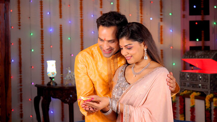 Indian husband-wife holding a well-lit clay Diya on the occasion of Diwali - Love and Bonding. Young man holding his wife while looking at an oil lamp burning brightly - festive season  Hindu festi...