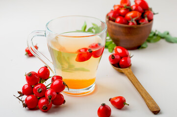 Cup with rosehip drink, bowl with rosehip berries, rosehip branches, berries in a wooden spoon on a light background, horizontal.