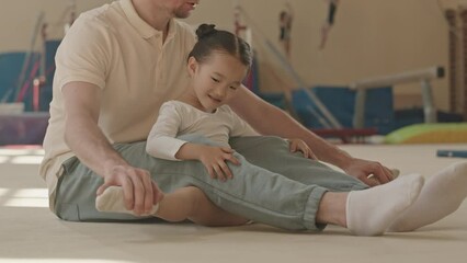 Professional Caucasian male gymnastics trainer helping 5 year old Asian girl with leg stretching during indoor class in spacious studio equipped with gymnastics facilities