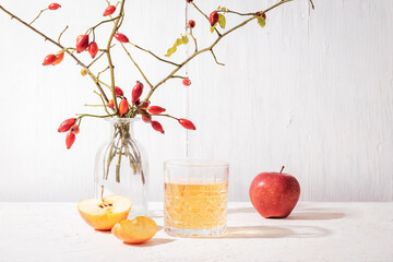 Fresh ripe apples and glass of apple juice or cider on white table. Vase with rosehip branches on...