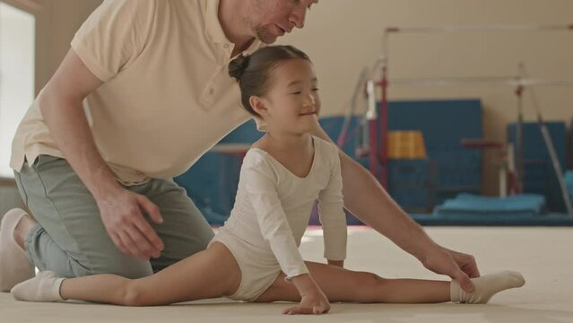 Skilled 5 year old Asian girl practicing splits while training with Caucasian male instructor in gymnastics class