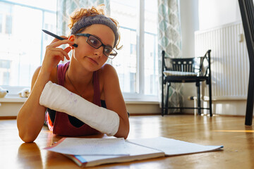 Teenage girl with broken arm in cast, wearing glasses, doing homework, at home