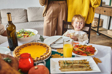 adorable toddler girl looking away and smiling near pumpkin pie during Thanksgiving celebration