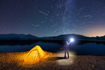A man with a lantern next to his camping tent is watching the Perseids meteor on the edge of a lake. Milky Way on the backgroung. Spain