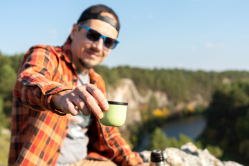 smiling man tourist in red checkered shirt drinking tea from thermos while sitting on rocky river bank in autumn and relaxing in nature