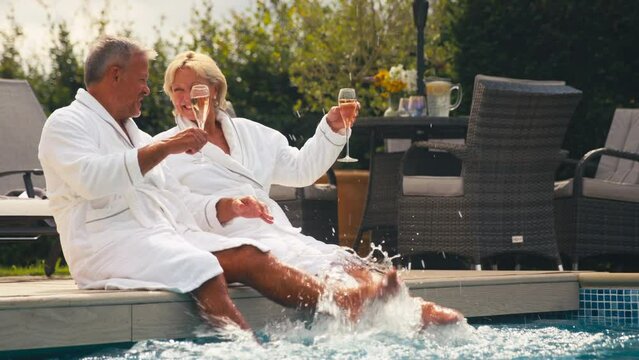 Senior retired couple wearing robes drinking champagne and splashing in hotel swimming pool on summer spa holiday - shot in slow motion