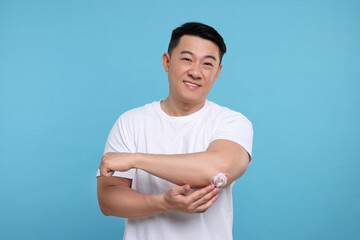 Handsome man applying body cream onto his elbow on light blue background