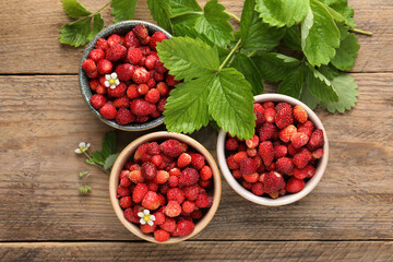Fresh wild strawberries in bowls and leaves on wooden table, flat lay