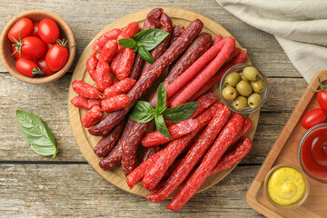 Different thin dry smoked sausages, basil, sauces and olives on wooden table, flat lay