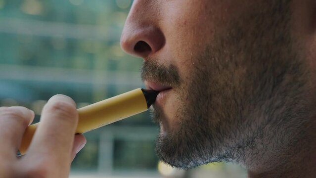 Close-up of young caucasian man with beard smoking an electronic cigarette outside office