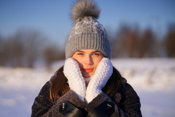 Portrait of beautiful girl young frozen pretty woman standing walking in winter snowy park at cold snow frosty day in hat, scarf, in gloves looking at camera