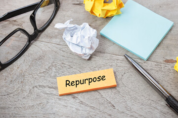 REPURPOSE word is written on the post-it note on the wooden table. A pen, crumpled papers,...