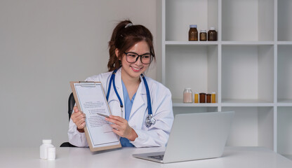 young asian lady doctor in white medical uniform with stethoscope using computer laptop talking...