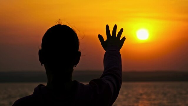 Silhouette female hand stretches out to the sunset, slow motion. Setting sun rays between fingers of open woman palm. Arm to the sun, faith in god. Concept dream, happiness, freedom, mental wellness.