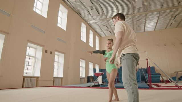Full length slowmo of little Caucasian girl in green leotard practicing front handspring with gymnastics coach while having training in spacious class