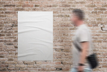 Wrinkled, blank A3 poster in white, adhered to a brick wall. A man walks beside. Ad or marketing...