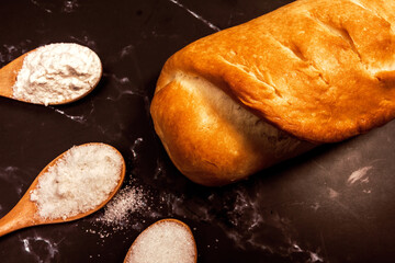 fresh homemade bread and three wooden spoons with sugar salt and flour on a black marble background