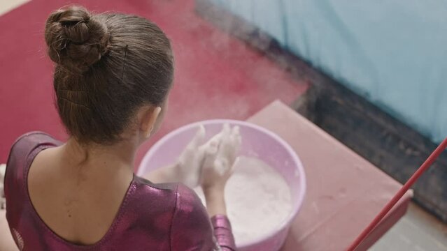 High angle slowmo shot of female gymnast in purple leotard using chalk powder on her hands for better bar grip