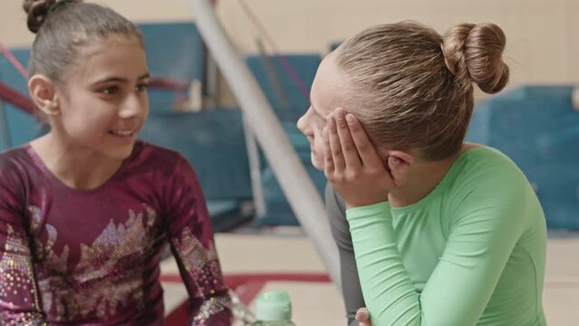 Tilt up of two diverse little girls sitting on gymnastics bean and having conversation while resting after group training in studio