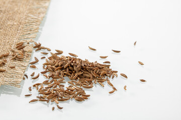A bunch of cumin seeds next to a burlap napkin on a white background