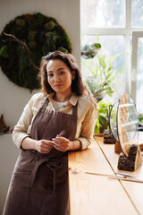 Portrait of asian craftswoman in apron looking at camera near framed herbariums on wooden stands in...