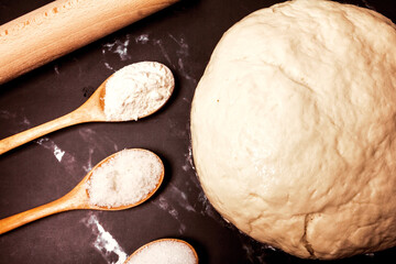 yeast dough, rolling pin and three wooden spoons with sugar salt and flour on a black marble background