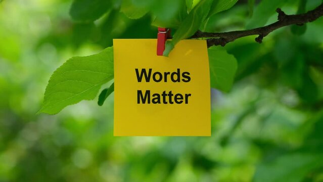A yellow paper note with the phrase Words Matter on it attached to a tree branch with a clothes pin. Close up.