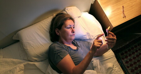 Woman picks up phone from nightstand lying in bed. Older lady browsing internet on smartphone before sleep