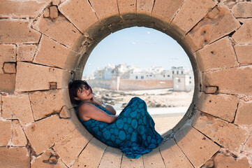 Attractive South American Girl Posing In A Resting Moment In A Circular Hole In A Fortress Wall While Traveling In Essaouira, Morocco.JPG - obrazy, fototapety, plakaty