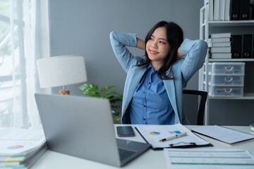 A young Asian businesswoman sits in a chair relieved from fatigue as she sits in the office with a happy smile at business success.